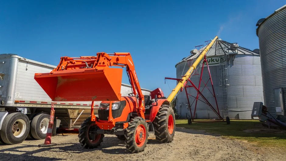 Bright orange tractor with front loader and grain auger near silos under clear sky