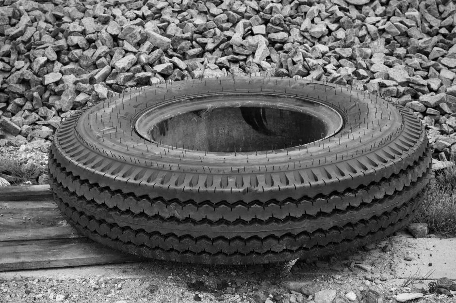 Black and white photo of a large industrial tire resting on gravel, showcasing rugged texture and urban setting