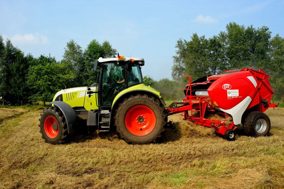 Green tractor and hay baler working on a farm field with trees in the background