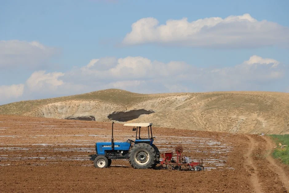 A blue tractor plowing a vast farmland against a backdrop of rolling hills and clouds