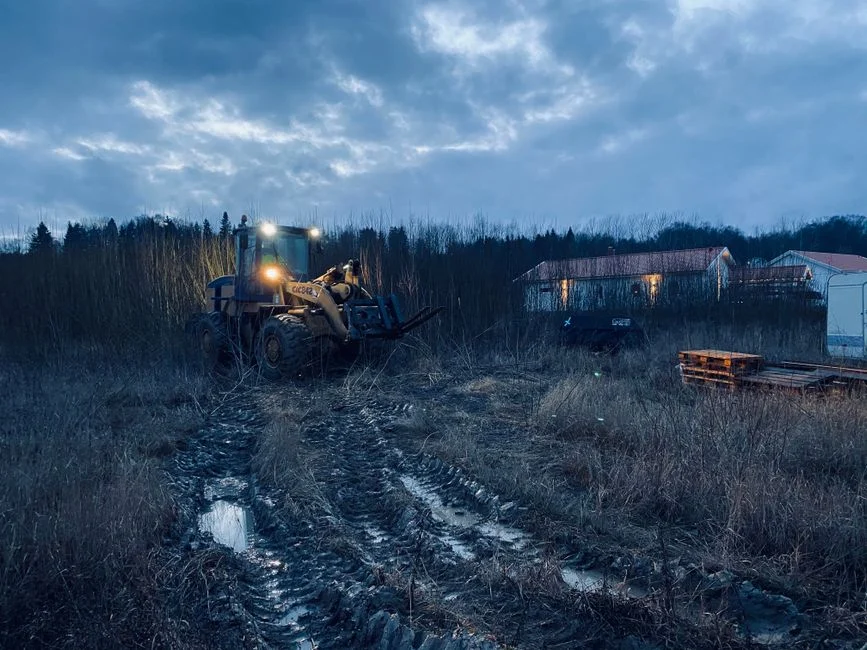 Tractor Tire Types Explained: R1 vs R2 vs R3 vs R4 Tractor working in a muddy, rural field at dusk, under a cloudy sky, near illuminated farm buildings
