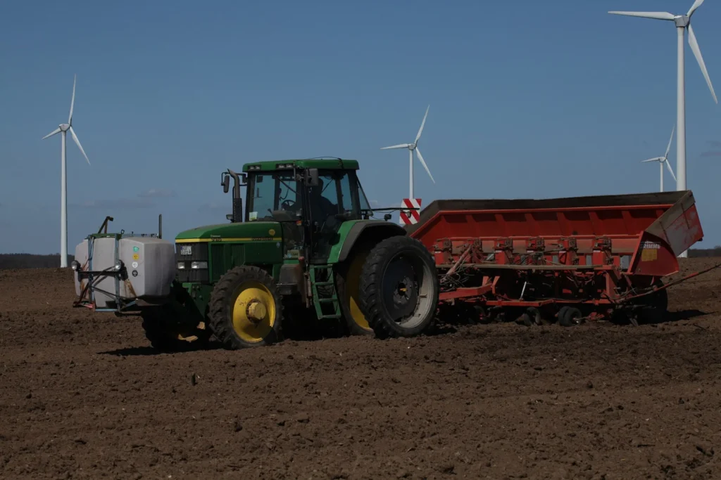 Green tractor pulling a planter across a dark field with wind turbines behind