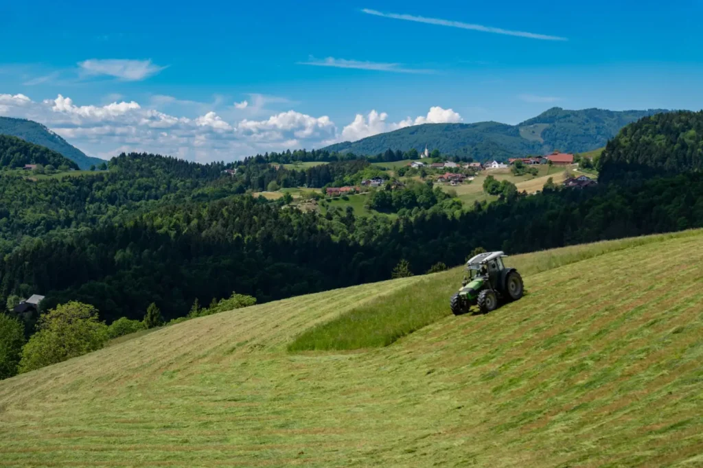 Tractor mowing grass on a green hillside with mountains in the distance