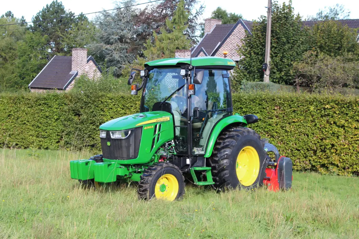 A green tractor mowing grass in a picturesque countryside landscape with houses and trees.