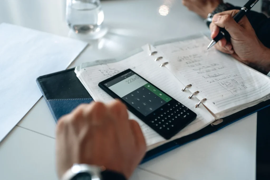 Hands writing in a notebook beside a phone calculator on a table