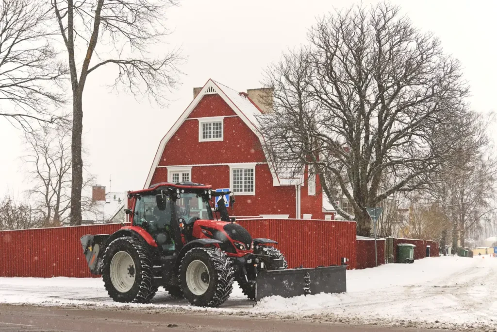 Red tractor plowing snow beside a red house