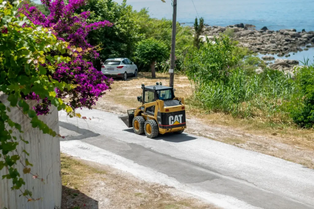 Skid steer loader parked on a sunny coastal road with green foliage and the sea behind