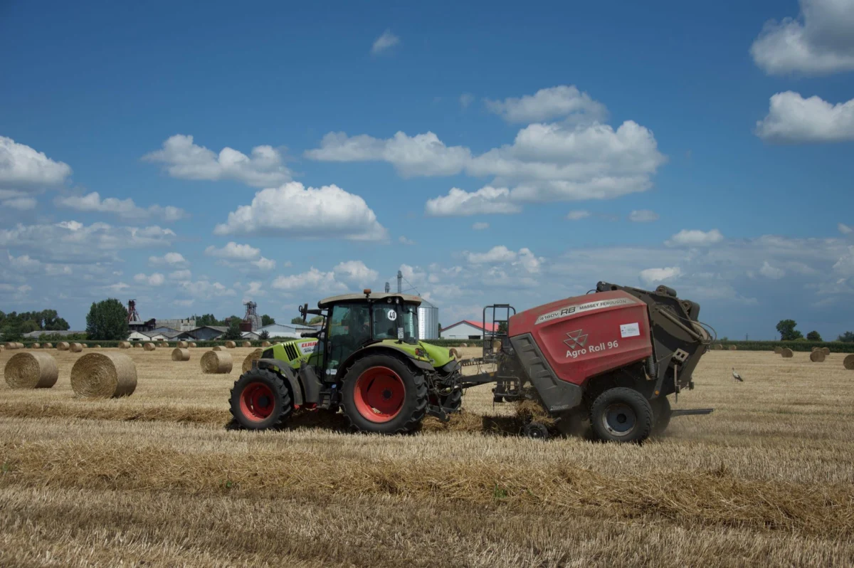 Tractor and baler working in a sunny field, surrounded by hay bales and buildings.