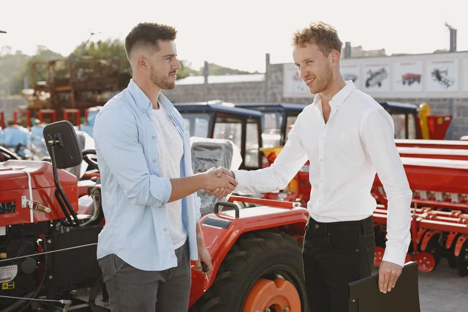 Two men shaking hands near a tractor, symbolizing a business deal or purchase agreement, outdoors