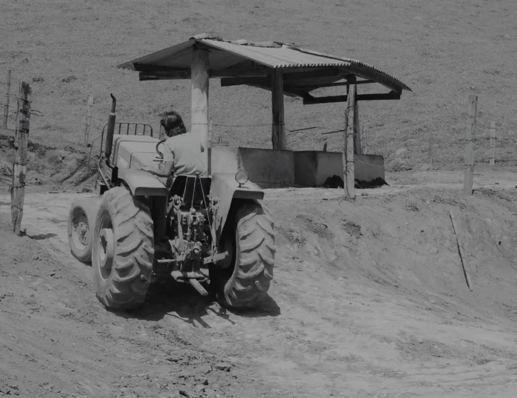 Back view of a farmer driving a tractor on a dirt path near a simple wooden shed.